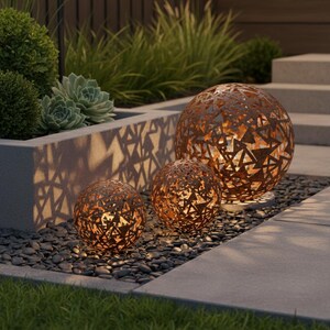 May include: Three decorative metal spheres with geometric cutouts, illuminated from within, creating a warm glow. The spheres are in varying sizes and have a rusted finish. They are placed on a bed of dark pebbles, with greenery in the background.