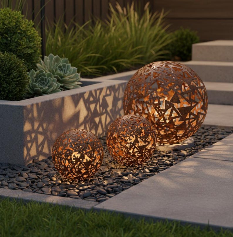 May include: Three decorative metal spheres with geometric cutouts, illuminated from within, creating a warm glow. The spheres are in varying sizes and have a rusted finish. They are placed on a bed of dark pebbles, with greenery in the background.