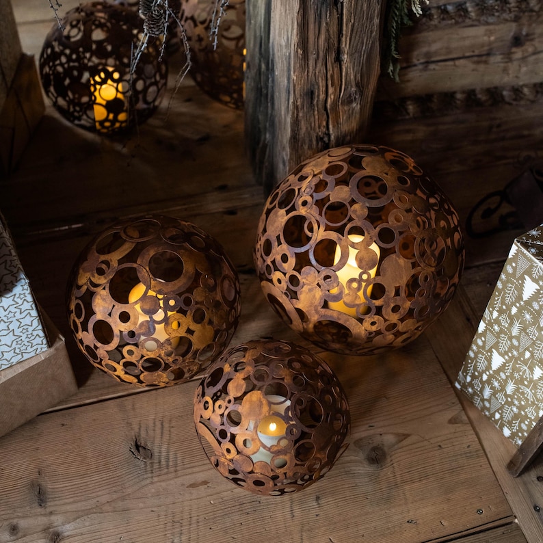 May include: Three spherical, metal candle holders with a rusted brown finish. Each sphere is intricately cut with various sized circular holes, allowing the warm candlelight within to glow. The candle holders are placed on a wooden surface.