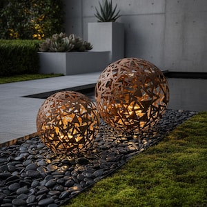 May include: Two spherical outdoor lights with a geometric design. The lights are made of a rusty-brown metal and sit on black pebbles. The lights are illuminated from within, casting a warm glow. The background includes greenery and a concrete wall.