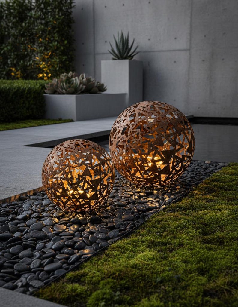 May include: Two spherical outdoor lights with a geometric design. The lights are made of a rusty-brown metal and sit on black pebbles. The lights are illuminated from within, casting a warm glow. The background includes greenery and a concrete wall.