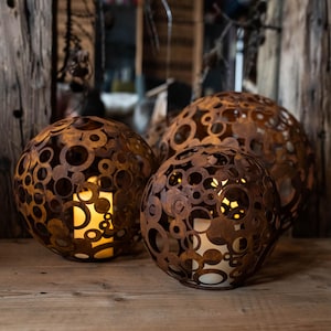 May include: Three spherical, rusty-brown metal lanterns with circular cutouts. Each lantern has a warm, glowing light inside, creating a cosy ambiance. The lanterns are of varying sizes and sit on a wooden surface, ideal for home decor.