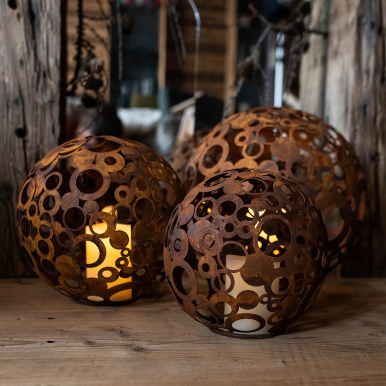 May include: Three spherical, rusty-brown metal lanterns with circular cutouts. Each lantern has a warm, glowing light inside, creating a cosy ambiance. The lanterns are of varying sizes and sit on a wooden surface, ideal for home decor.