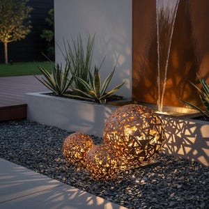 May include: Three decorative metal spheres with geometric cutouts, lit from within, rest on grey gravel. The spheres vary in size and emit a warm glow. The scene includes a water feature and greenery, creating a tranquil outdoor ambiance.
