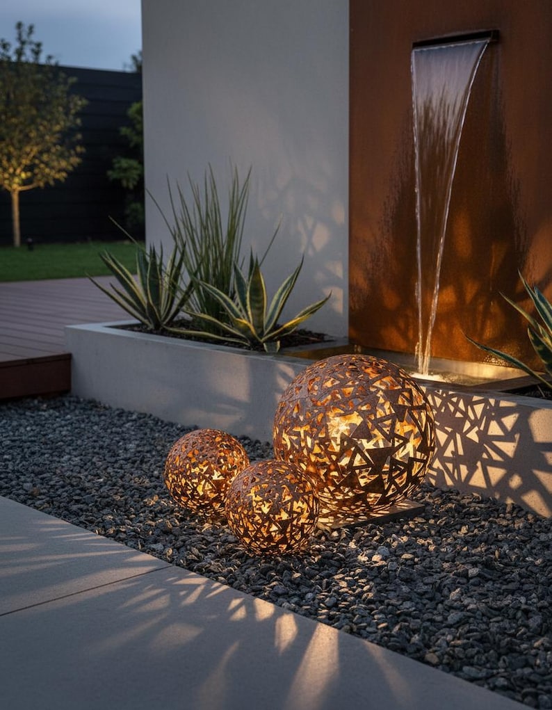 May include: Three decorative metal spheres with geometric cutouts, lit from within, rest on grey gravel. The spheres vary in size and emit a warm glow. The scene includes a water feature and greenery, creating a tranquil outdoor ambiance.