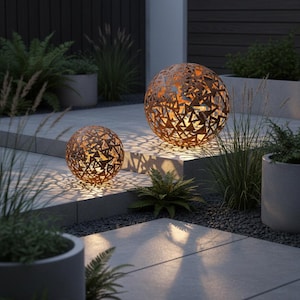 May include: Two decorative metal spheres with geometric cutouts, lit from within, creating patterned shadows. The spheres are on a stone patio, surrounded by plants, creating a warm outdoor atmosphere. The spheres are a rusty brown colour.