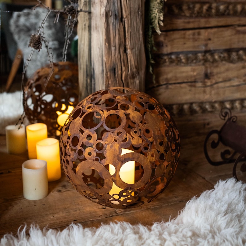 May include: A spherical, rusty-brown metal lantern with a pattern of various-sized circles. The lantern is lit from within, casting a warm glow. Several unlit, cream-coloured candles and a second, similar lantern are in the background, suggesting a rustic, cosy theme.