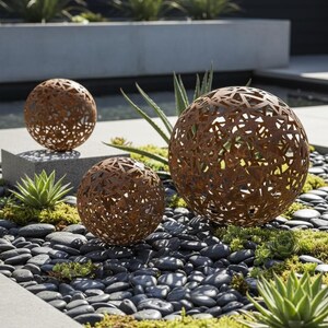 May include: Three decorative metal spheres with intricate cut-out patterns in a garden. The spheres have a rusted brown finish and vary in size. They are set amongst black pebbles, succulents, and greenery, creating a modern outdoor aesthetic. The spheres are made of metal.