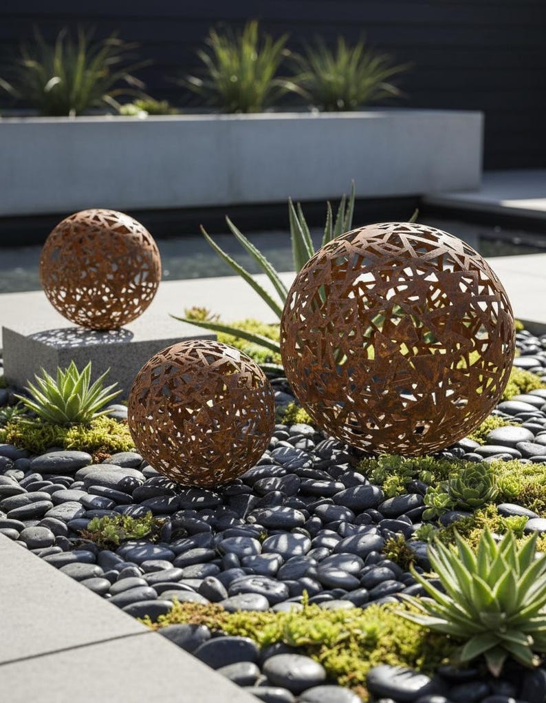 May include: Three decorative metal spheres with intricate cut-out patterns in a garden. The spheres have a rusted brown finish and vary in size. They are set amongst black pebbles, succulents, and greenery, creating a modern outdoor aesthetic. The spheres are made of metal.