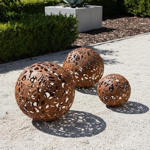 Op de afbeelding: Drie decoratieve, bolvormige metalen tuinornamenten met ingewikkelde uitgesneden patronen. De bollen, in verschillende maten, hebben een roestbruine afwerking. Ze staan op een lichtgekleurd grindoppervlak, met groen en een betonnen plantenbak op de achtergrond.