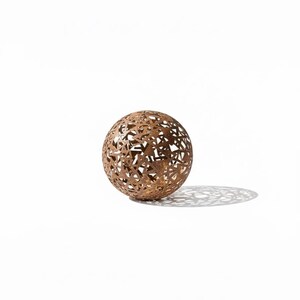 May include: A decorative metal sphere with a geometric openwork design. The sphere is a warm brown colour and casts a patterned shadow on the white surface. The sphere is a decorative item.