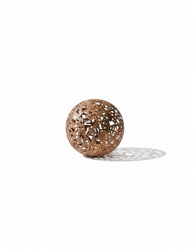 May include: A decorative metal sphere with a geometric openwork design. The sphere is a warm brown colour and casts a patterned shadow on the white surface. The sphere is a decorative item.