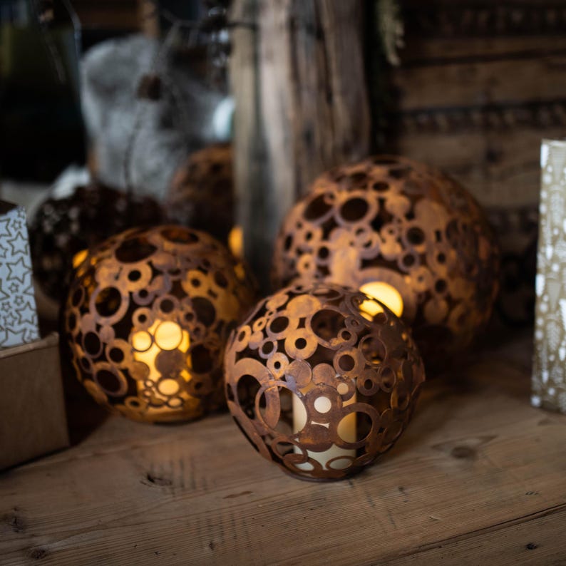 May include: Three spherical, rusty-brown metal lanterns with circular cutouts, each housing a warm, glowing light. The lanterns vary in size and are arranged on a wooden surface, creating a cosy, ambient display. The design suggests a rustic, decorative theme.
