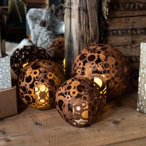 May include: Three spherical, rusty-brown metal lanterns with circular cutouts, each housing a warm, glowing light. The lanterns vary in size and are arranged on a wooden surface, creating a cosy, ambient display. The background includes wooden elements and decorative accents.