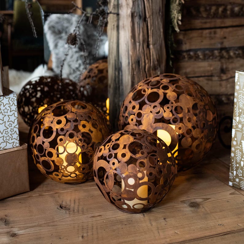 May include: Three spherical, rusty-brown metal lanterns with circular cutouts, each housing a warm, glowing light. The lanterns vary in size and are arranged on a wooden surface, creating a cosy, ambient display. The background includes wooden elements and decorative accents.