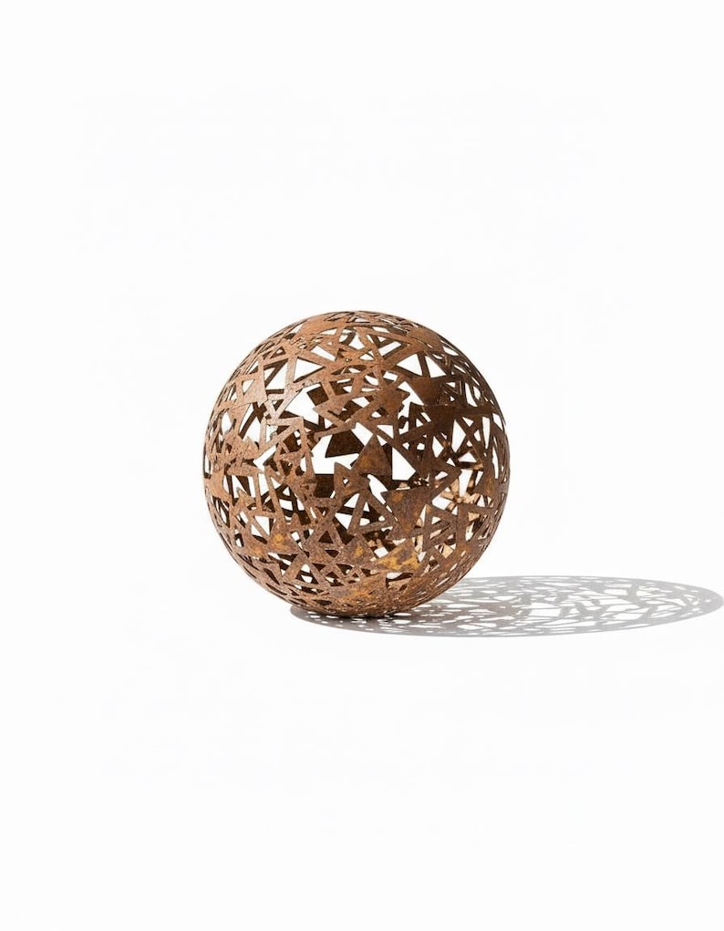 May include: A decorative metal sphere with a rusted brown finish. The sphere features a geometric pattern of cut-out shapes, creating an openwork design. The sphere casts a shadow on the white surface.