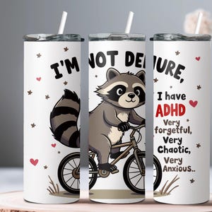 May include: White tumbler featuring a cartoon raccoon riding a bicycle. The tumbler has the text "I'm not deure, I have ADHD Very forgetful, Very Chaotic, Very Anxious..." printed on it.