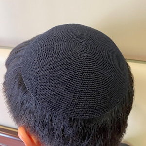 May include: A black, circular kippah, a traditional Jewish head covering, is shown from a slightly elevated angle. The kippah is made of a tightly woven, dark fabric, with a spiral pattern emanating from the center. The person's dark hair and ear are visible.