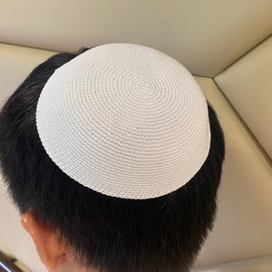 Può includere: Una kippah bianca all'uncinetto, o yarmulke, è appoggiata su capelli scuri. Il berretto circolare è realizzato con filato di colore chiaro, a trama fitta. L'immagine è ripresa da un'angolazione leggermente rialzata, mostrando la parte posteriore della testa e la parte superiore del berretto.