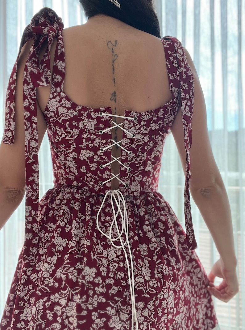 May include: A red and white floral dress with a lace-up back and tie straps. The dress has a fitted bodice and a flowy skirt.