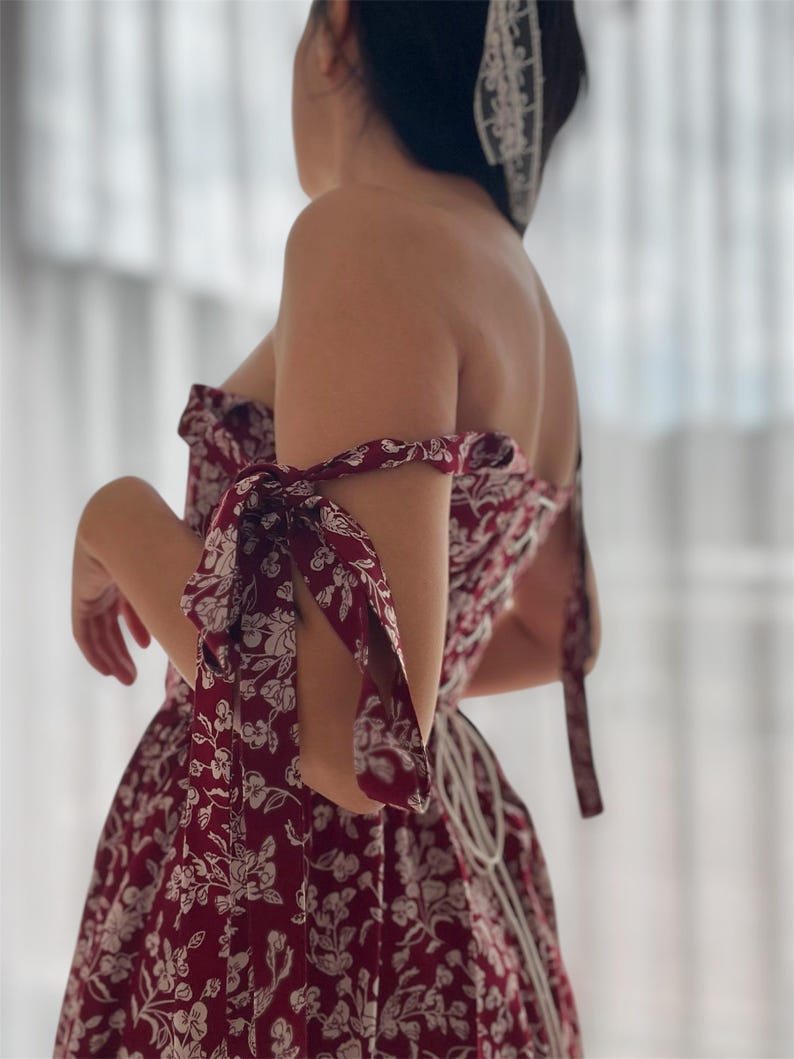 May include: A red and white floral print dress with a fitted bodice and a full skirt. The dress has a sweetheart neckline and is strapless. The sleeves are tied at the shoulders with matching fabric.