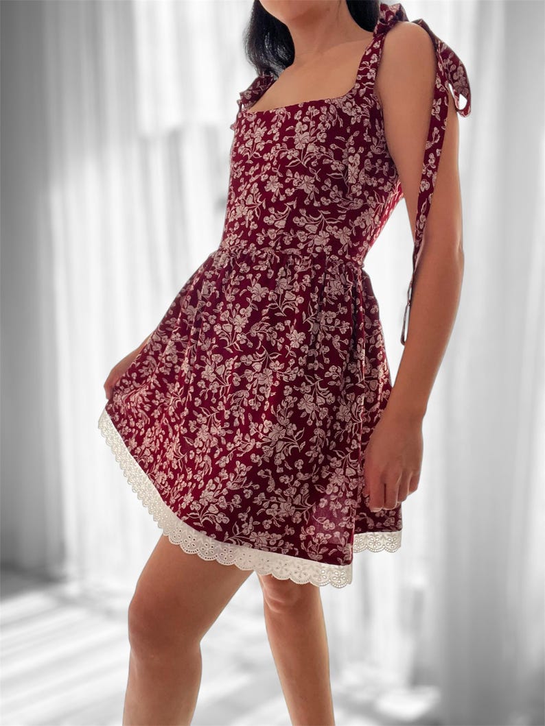 May include: A burgundy floral print dress with a square neckline and thin straps. The dress has a white lace trim around the hem and ties at the shoulders.