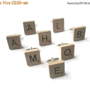May include: A collection of wooden square cufflinks, each displaying a letter and a number. The letters include A, H, M, E, L, B, and O. The numbers are 1, 3, and 4. The cufflinks are a light brown colour and are on a white surface.