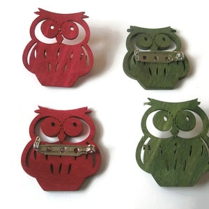 May include: Four wooden owl brooches, two red and two green. The owls have large eyes and a detailed design. Each brooch has a metal pin on the back.