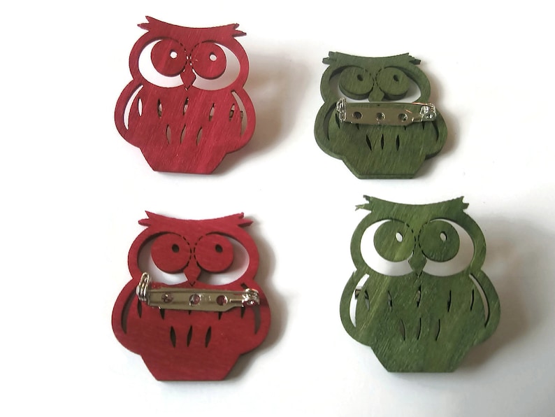May include: Four wooden owl brooches, two red and two green. The owls have large eyes and a detailed design. Each brooch has a metal pin on the back.