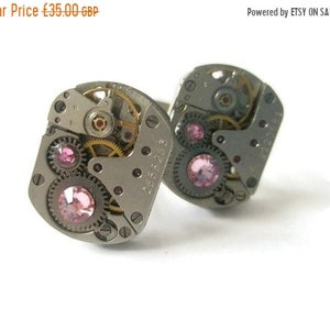 May include: A pair of silver cufflinks with pink rhinestones set in a watch movement design. The cufflinks feature a silver gear and a silver plate with the number "2566218".