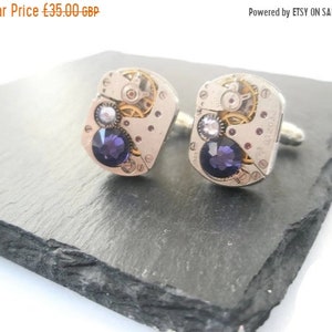May include: A pair of silver cufflinks featuring a purple and clear crystal design. The cufflinks are crafted from watch parts and have a steampunk aesthetic.
