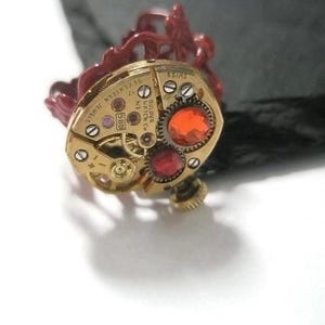 May include: A gold-toned watch movement with red and orange jewels, set in a red plastic ring. The watch movement has the text "BULOVA WATCH CO. 17 JEWELS 5-1114145" printed on it.