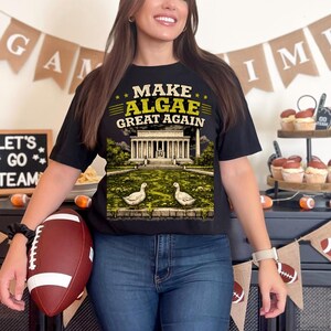 May include: A black t-shirt featuring a graphic print with the text "MAKE ALGAE GREAT AGAIN" above an illustration of the Lincoln Memorial and a reflecting pool filled with green algae and two white ducks standing on the edge of the water.