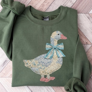 May include: An olive green sweatshirt featuring a goose applique. The goose has a blue and white patterned body, a red and white checkered beak and feet, and a blue and white plaid bow. The sleeves have a gathered cuff.