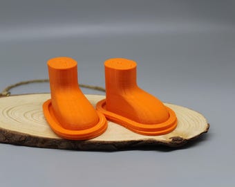 May include: A pair of bright orange, 3D-printed boot-shaped objects with smooth, rounded toes and cylindrical shafts. The boots feature a distinct layered texture from the printing process and are set on a flat, oval-shaped base.