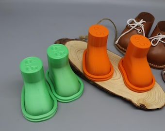 May include: Two pairs of 3D printed boot-shaped shoe stretchers in bright green and orange, each embossed with the number 6.5 on the top. The stretchers are arranged on a natural wood slice, positioned near two small brown leather shoes with white laces.