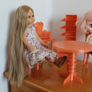 May include: Two dolls are seated near an orange table and chairs. The doll on the left has long blonde hair and wears a floral dress and brown boots. The doll on the right has pink hair and wears a floral top and black shorts.