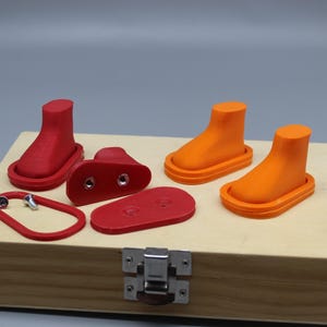 Puede incluir: Un conjunto de componentes con forma de bota impresos en 3D en rojo y naranja, que incluye dos botas rojas ensambladas, dos botas naranjas ensambladas, dos placas base rojas planas, un marco rojo y dos pequeños tornillos metálicos sobre una caja de madera.