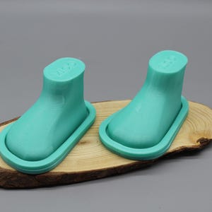 Könnte beinhalten: Zwei türkisblaue Plastik-Schuhformen auf einer Holzoberfläche. Die Formen sind bootförmig und haben eine abgerundete Spitze.
