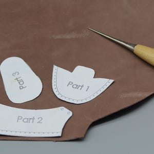 Puede incluir: Tres patrones de papel para un proyecto de cuero, etiquetados como "Parte 1", "Parte 2" y "Parte 3", se colocan sobre una superficie de cuero marrón. También hay un punzón de cuero con mango de madera sobre la superficie.