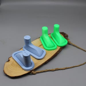 May include: Three 3D-printed shoe-shaped objects in light blue and green, displayed on a wooden slice with a jute rope. The objects have a rectangular base with a rounded top and a cylindrical post.