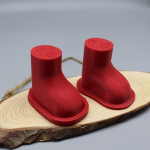 以下が含まれることがあります： 2つの赤い3Dプリントされたブーツ型のオブジェクト。各オブジェクトの上部に「6.0」と印刷されています。オブジェクトは木製の表面の上に置かれています。