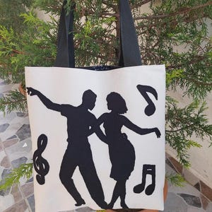 May include: White canvas tote bag with black handles. The bag features a black silhouette of a couple dancing with musical notes.
