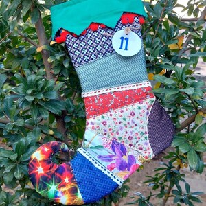 May include: A patchwork Christmas stocking with a green top and a white circle with the number 11 on it. The stocking is made of different fabrics with various patterns, including floral, geometric, and paisley.