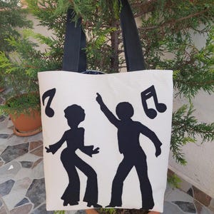 May include: A white canvas tote bag with black handles. The bag features a black silhouette of two people dancing with musical notes above them.