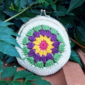 May include: A round, crocheted coin purse with a metal clasp. The purse features a colorful floral design in the center, with pink, yellow, purple, and green petals. The purse is set against a backdrop of green leaves and a brick surface.