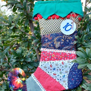 May include: A colorful patchwork Christmas stocking with a green cuff and red pom-poms. The stocking has a blue and white paisley pattern, red and white polka dots, and a butterfly print. The stocking has a number "10" on a white circle tag.