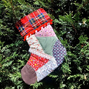May include: A patchwork Christmas stocking with a red plaid top, pink and white floral fabric, green fabric, and purple fabric with a white floral pattern. The stocking has a white lace trim and red pom-poms.