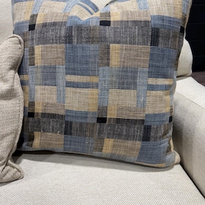 May include: Square decorative pillow with a geometric pattern in shades of blue, tan, and black. The pillow is placed on a light-colored sofa, showcasing its textured fabric and design.