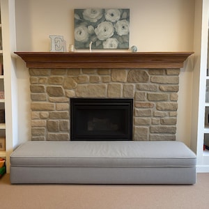 May include: A long, rectangular light gray upholstered bench with piped edges positioned in front of a stone fireplace. The fireplace features a dark metal insert, a wooden mantel, and a floral painting with white roses on a gray background.